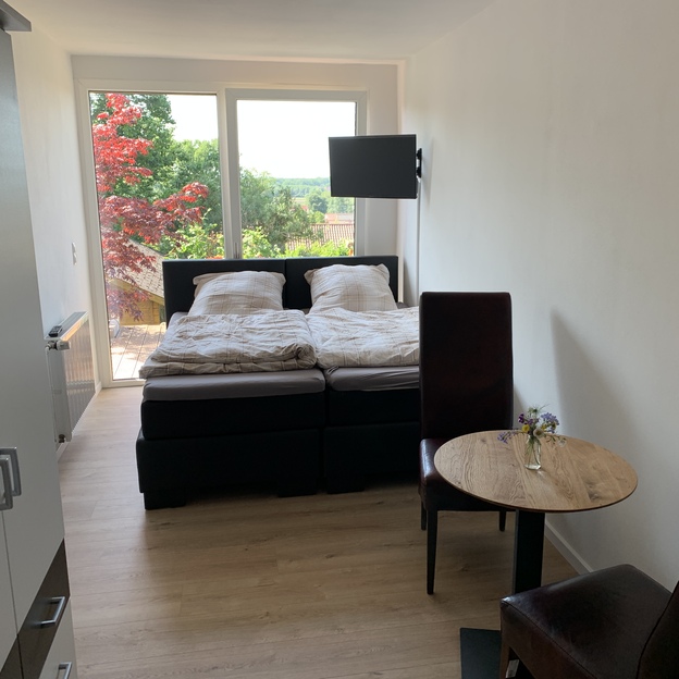 Doppelbett vor Fensterfront hinter zwei Stühlen und kleinem Tisch