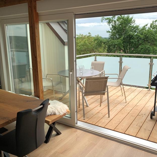 Blick durch eine offene Schiebetür eines Fensters nach Maß auf eine Holzterrasse mit Möbeln und Grill, umgeben von grüner Landschaft.