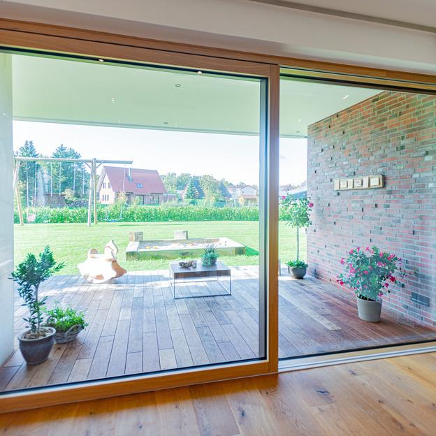 Ein heller Raum mit einer großen Schiebetür eines Fensters nach Maß, der zu einer Holzterrasse und einem grünen Garten führt.