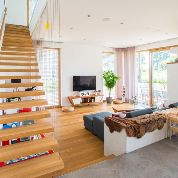 Einladendes Wohnzimmer mit Holzböden und -treppen, gemütlichem Sofa und großen Fenstern nach Maß mit Blick ins Grüne.