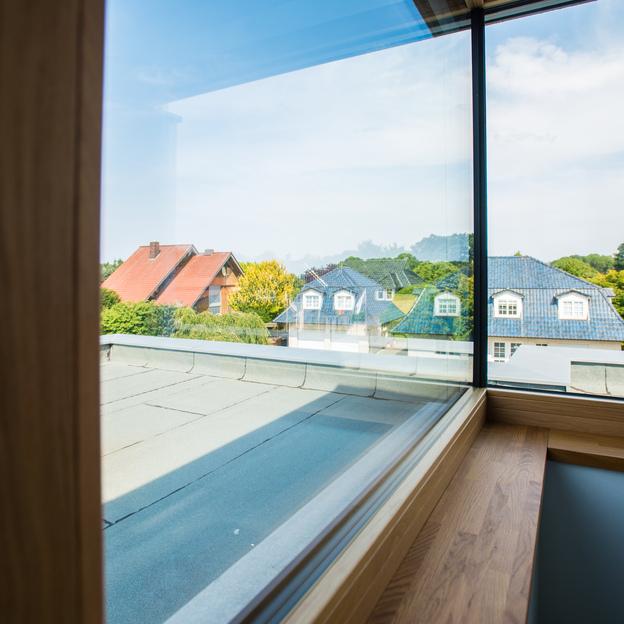 Innensicht auf ein bodentiefes Ganzglaseckfenster nach Maß mit Ausblick auf idyllische Hausdächer und grüne Bäume.