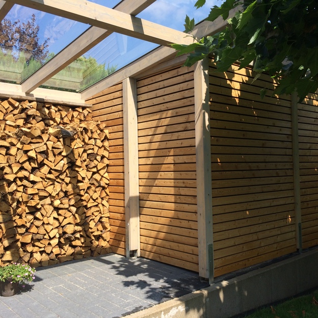 Gartenhaus mit Glasdach und Holzverschalung, daneben offenes Holzlager mit aufgestapeltem Brennholz auf gepflastertem Boden.