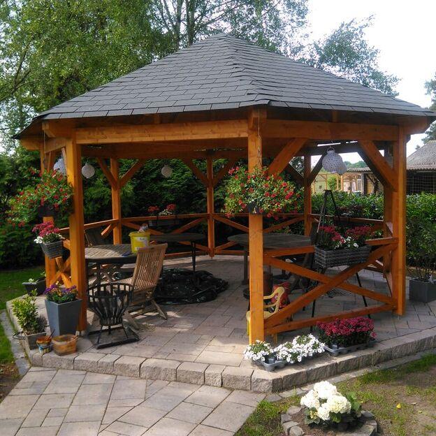 Achtseitiges offenes Gartenhaus aus Holz mit Schindeldach, Sitzmöbeln, Blumen und gepflastertem Boden im grünen Garten.