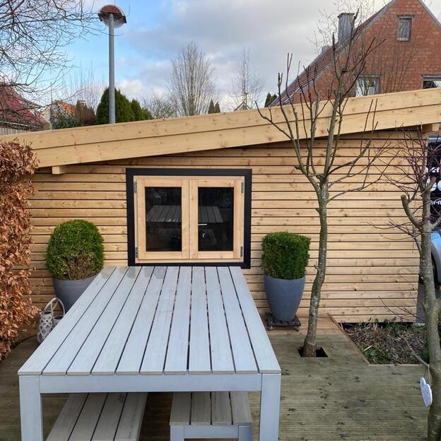 Modernes Gartenhaus mit Holzverkleidung und Flachdach, mittig ein Fenster mit schwarzem Rahmen, davor Esstisch mit Bank.