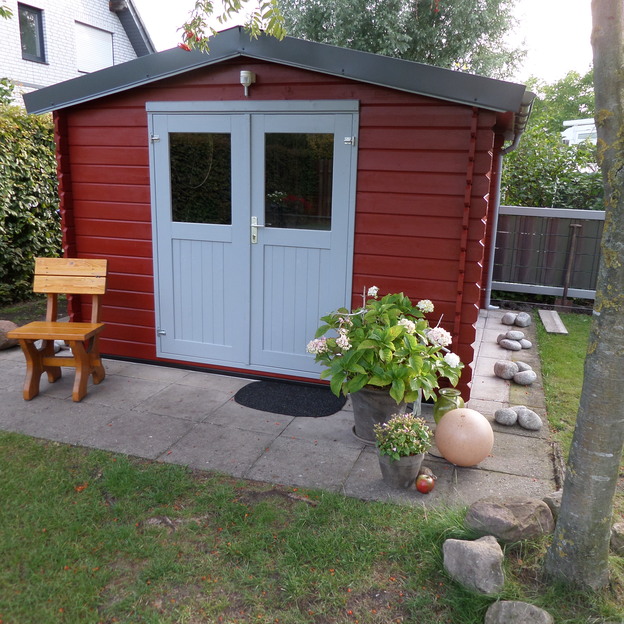 Gartenhaus mit rotem Holzanstrich und grauer Tür, davor Sitzbank, Blumenkübel und dekorativer Weg aus Natursteinen.