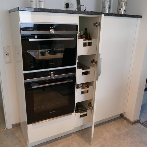 Offener Schrank einer Küche nach Maß zeigt Regale mit Lebensmitteln neben Einbauöfen, auf einer weißen und grauen Farbpalette.