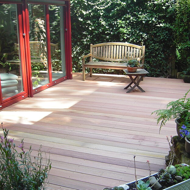 Holz-Terrassendielen vor roter Glasfassade, mit Holzbank, Beistelltisch und Blumentöpfen in grünem Gartenumfeld.