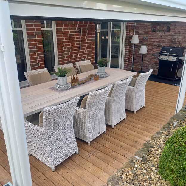 Terrassendielen aus Holz unter Glasüberdachung, mit großem Esstisch und weißen Rattanstühlen an einem roten Klinkerhaus.