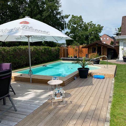 Terrassendielen aus Holz umrahmen einen eingelassenen Pool mit Sonnenschirm, Sitzmöbeln und Gartenhaus im Hintergrund.