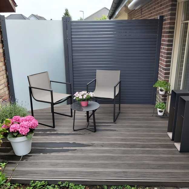 Kleine Terrasse mit dunklen Terrassendielen, zwei Stühlen, Blumentöpfen und modernem Sichtschutz aus Glas und Metall.