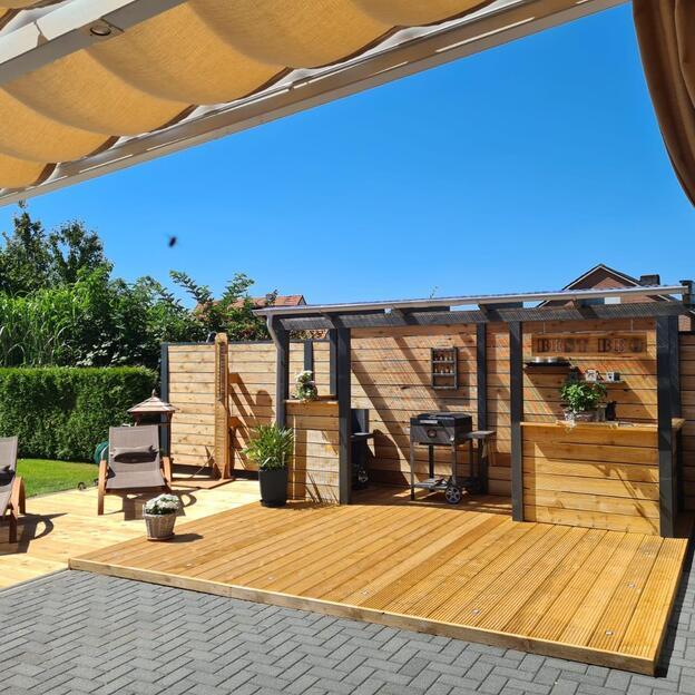 Terrassendielen aus Holz mit integrierter Outdoor-Küche, Grillplatz, Sonnenliegen und Sichtschutz im modernen Garten.