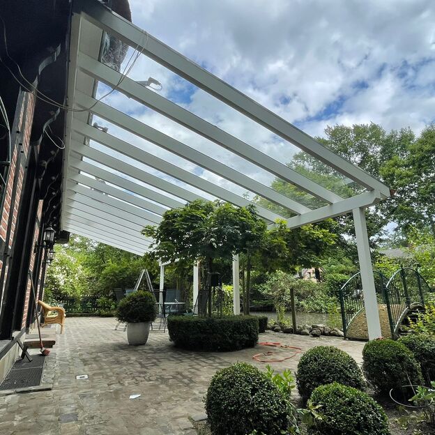 Weiße Terrassenüberdachung aus Glas und Aluminium an einem Backsteinhaus, mit Blick auf Garten und Sitzbereich.