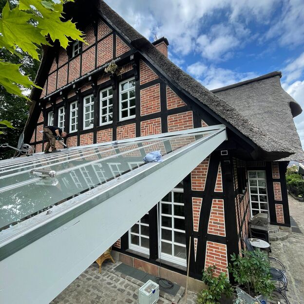 Montage einer Terrassenüberdachung mit Glasdach an einem rot-schwarzen Fachwerkhaus mit Reetdach und Kopfsteinpflaster.