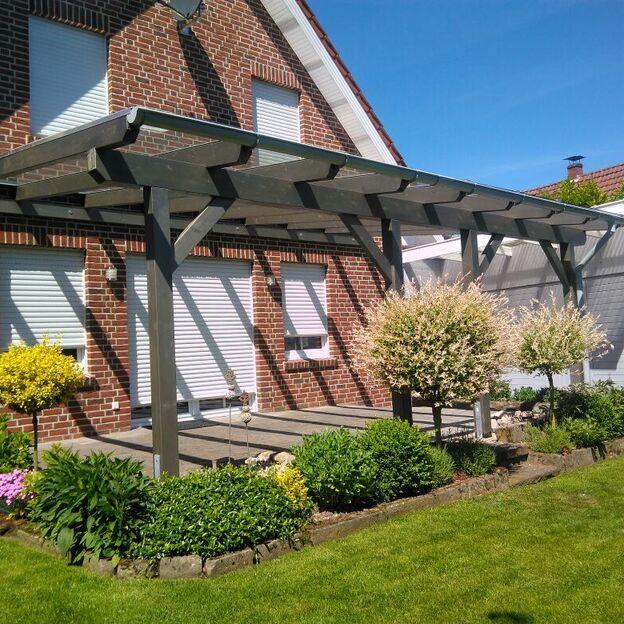 Terrassenüberdachung mit anthrazitfarbenem Gestell an einem roten Klinkerhaus, umgeben von gepflegten Beeten und Rasen.