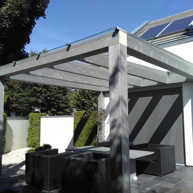 Terrassenüberdachung aus grauem Holz mit Glasdach an einem modernen Haus, darunter Sitzgruppe auf dunklen Bodenplatten.