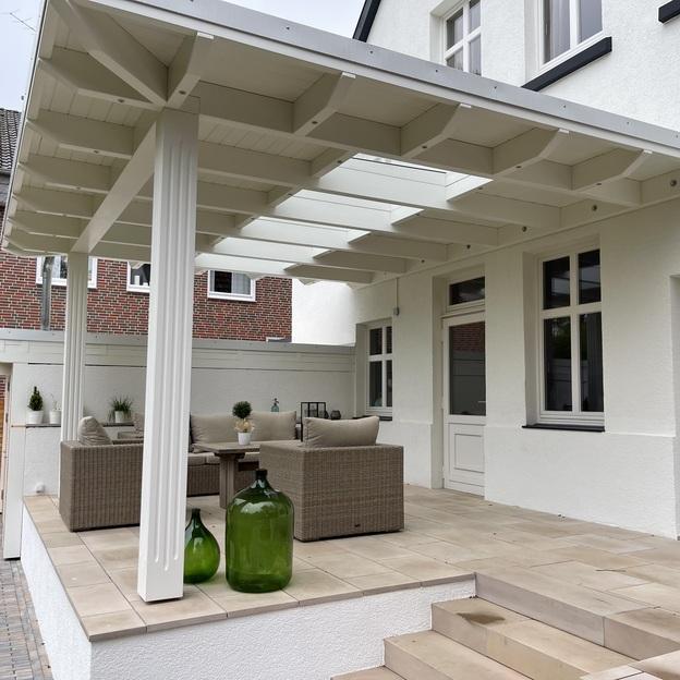 Weiße Terrassenüberdachung im klassischen Stil mit verzierten Pfosten, darunter gemütliche Sitzgruppe auf erhöhter Terrasse.