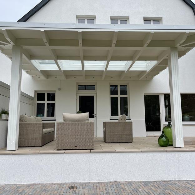 Weiße Terrassenüberdachung mit Glaseinsatz über einer erhöhten Sitzterrasse an einem modernen, hellen Wohnhaus.