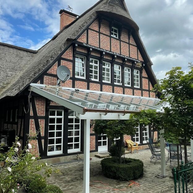 Weiße Terrassenüberdachung mit Glasdach an einem historischen Fachwerkhaus mit Reetdach und Kopfsteinpflaster-Terrasse.