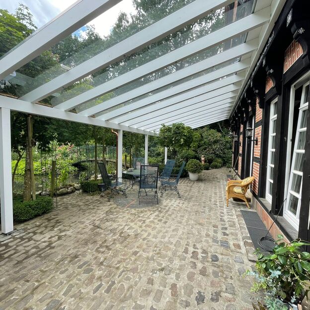Terrassenüberdachung mit Glasdach an einem Fachwerkhaus, darunter Sitzgruppe mit Blick auf Teich und Gartenbrücke.