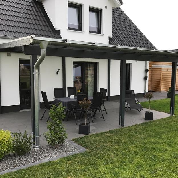 Terrassenüberdachung mit dunklem Gestell und klarer Dacheindeckung an einem weißen Einfamilienhaus mit Gartenmöbeln.