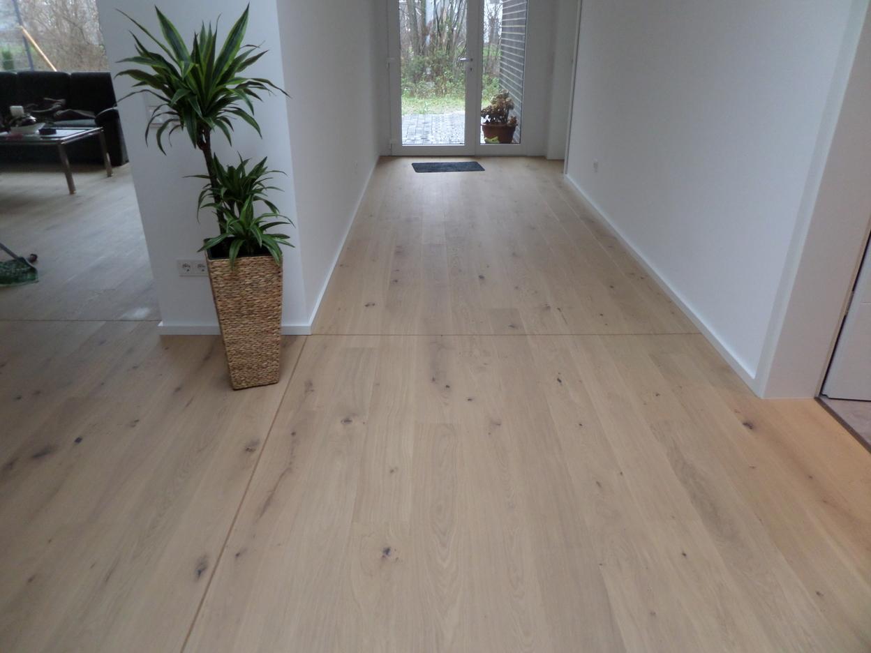 Langer, heller Flur mit breiten Eichenholzdielen, weißen Wänden und einer hohen Zimmerpflanze im geflochtenen Topf.