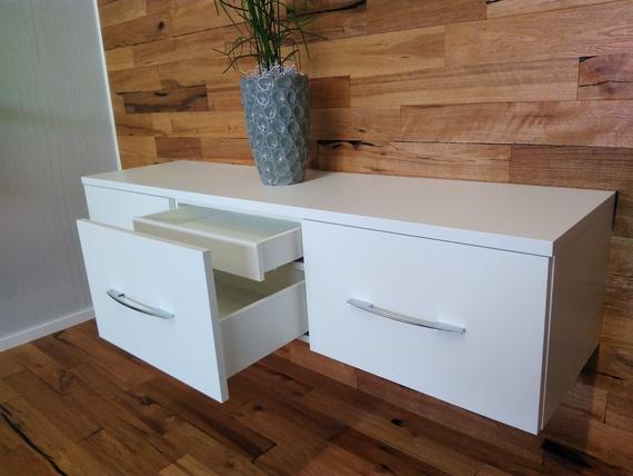 Modernes, weißes Hängesideboard mit geöffneten Schubladen vor einer Wand aus rustikalen Holzpaneelen.