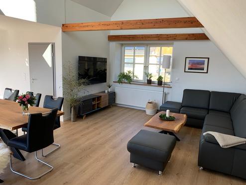 Freundlich eingerichtetes Wohnzimmer mit schwarzem Ledersofa, Holzboden und offenem Essbereich mit Holztisch.